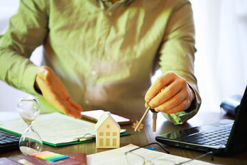 Real Estate Agent Holding House Keys Over Model Home at Desk With Paperwork and Laptop