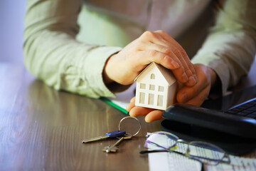 Hands Protecting Model House on Desk with Keys and Documents