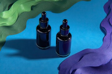 Two dark glass dropper bottles sit on a vibrant blue surface, partially obscured by sculpted green and purple fabric waves casting shadows