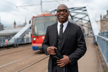 Man in formal attire stands confidently on a bridge with a tram approaching in an urban setting...