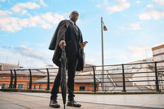 Businessman in formal attire standing on city street with an umbrella while checking his phone under cloudy sky