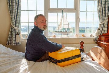 Individual sits on bed with yellow suitcase near window looking out at ocean during daytime in...