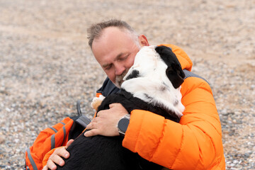 Man embraces black and white dog on beach during calm afternoon, capturing a moment of joy and...