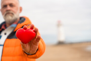 Man holds red heart symbol at beach with lighthouse in background during overcast day