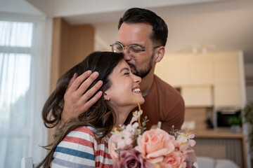 Man kissing woman on forehead and giving flowers bouquet at home