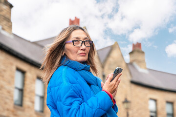 Person in blue jacket and scarf holding smartphone outdoors in a historical urban area with cloudy...