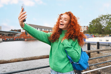 Woman with curly red hair takes a selfie by the riverside in spring while wearing a green sweater...