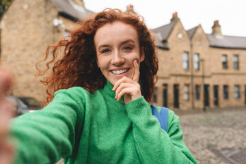 Woman with curly red hair poses for a selfie on a cobblestone street in a quaint neighborhood...