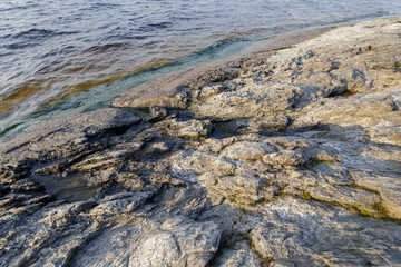 Rough, weathered rock formations meet the edge of a calm lake, with small pools and algae lining the shoreline.