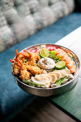 Grilled shrimp, salmon slices, dumplings, and dipping sauce beautifully arranged in a metal bowl with greens and lime halves — a vibrant gourmet seafood platter on white stones.