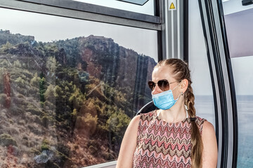 Woman wearing a face mask and sunglasses enjoying a scenic ride on the Alanya Teleferik cable car. The background reveals a mountainous landscape and distant sea views