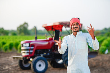 young indian farmer standing with tractor and showing smartphone at field