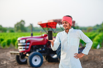young indian farmer standing with tractor and showing smartphone at field