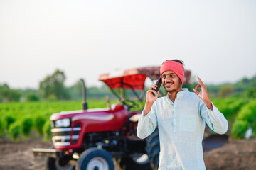 indian farmer standing with tractor and talking on smartphone at field