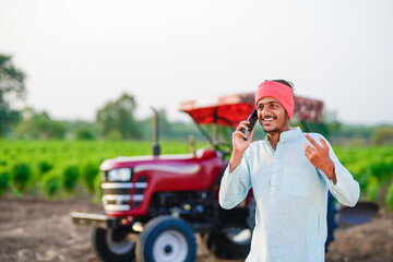 indian farmer standing with tractor and talking on smartphone at field