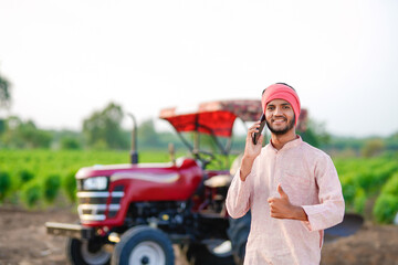 indian farmer standing with tractor and talking on smartphone at field