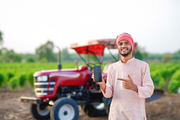 young indian farmer standing with tractor and showing smartphone at field