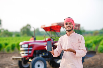 young indian farmer standing with tractor and showing smartphone at field