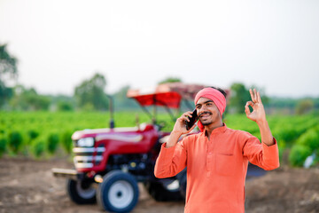 indian farmer standing with tractor and talking on smartphone at field