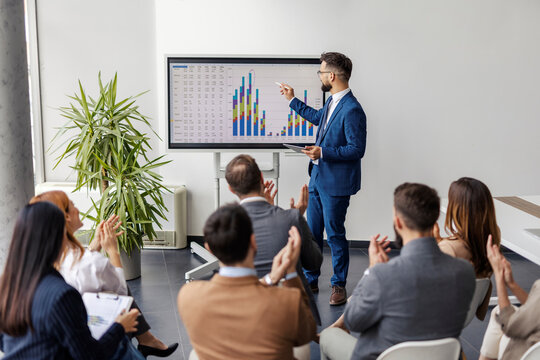Financial analyst is standing at conference room and presenting statistics with charts and numbers on interactive board in front of audience who is applauding.
