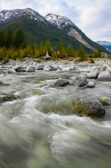 Flowing river amid rocky landscape with mountains and trees in the background on a cloudy day