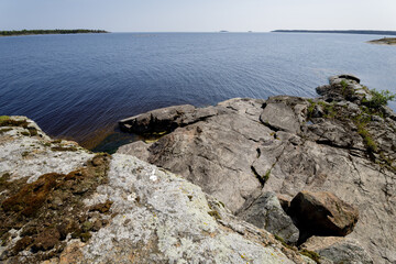 Granite rocks slope into a serene blue lake under a clear sky, with distant islands visible on the...