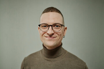 Portrait of young adult Caucasian man with short hair, wearing glasses and nose ring, smiling while looking directly at camera against plain background