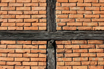 Orange-red brick wall features two intersecting weathered wooden beams forming a cross pattern,...