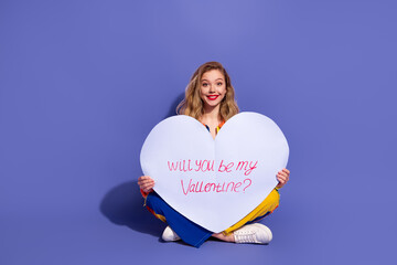 Smiling young woman holding a large heart sign with a romantic message, will you be my Valentine