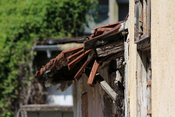 Close-up of an old building’s deteriorating roof, showing exposed wooden beams, broken red tiles, and weathered wall, with greenery blurred in the background