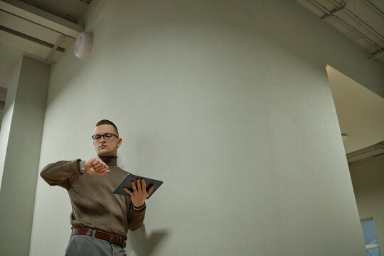 Caucasian young adult man standing indoors checking time on wristwatch while holding digital tablet in other hand, wearing glasses and looking slightly to side