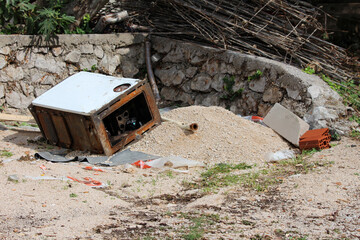 Rusted metal appliance lies on its side among gravel, bricks, and debris at a construction site,...