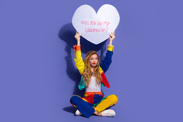 Casual young woman posing with a heart prop displaying Valentine's Day message will you be my...