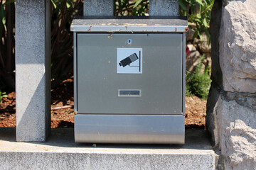Rectangular gray metal mailbox with a surveillance camera symbol sticker is mounted between stone and concrete posts, set in front of a garden with soil and foliage