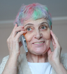 Happy elderly woman with multi-colored hair talking on cellphone, making call from home, speaking...