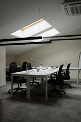 Empty modern office workspace featuring several black office chairs surrounding desks under skylight, various office supplies and coffee cups scattered on tables, no people present