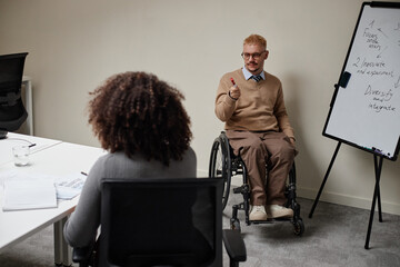 Caucasian young adult man with disability sitting in wheelchair leading business meeting, holding...