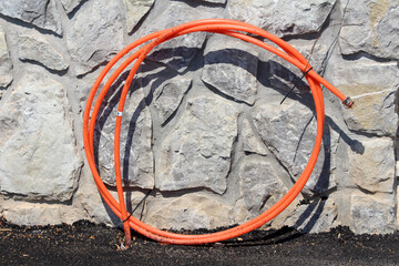 Bright orange flexible conduit pipe neatly coiled and fastened with wire, leaning against a textured stone wall, with black soil at the base in strong sunlight