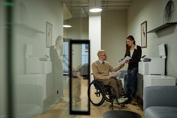 Wide shot of young Caucasian man with disability discussing documents with female colleague at...