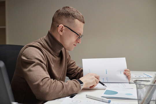 Caucasian young adult man analyzing financial documents at desk, holding paper with charts and graphs, wearing glasses, sitting in modern office environment, focused on work