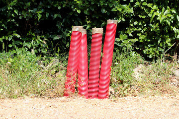Four bundles of red corrugated plastic pipes stand upright on the ground beside a dirt road, with dense green shrubs and wild grass in the background, ready for utility installation