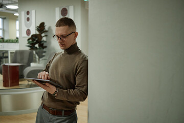 Caucasian young adult man standing in modern office leaning against wall using digital tablet with...