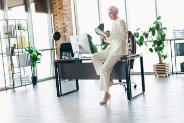 Mature businesswoman in formal attire sitting on office desk reading a tablet in a modern corporate...
