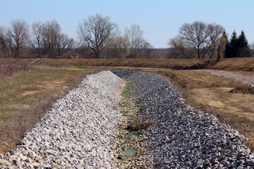 Wide drainage ditch lined with angular riprap stones runs through a cleared field, with sparse...