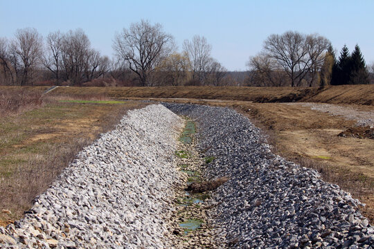Wide drainage ditch lined with angular riprap stones runs through a cleared field, with sparse grass and bare deciduous trees in the background under a clear blue sky