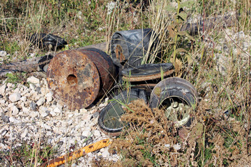 Pile of rusted metal drums and cracked rubber tires lies abandoned among white stones, dry grass,...