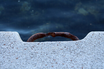 Close-up of a rusted metal mooring ring embedded in a textured concrete dock, with deep blue water...