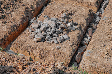 An outdoor construction site shows freshly dug trenches filled with rocks and gravel, marking the...