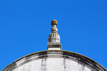 Ornate stone finial with weathered details sits atop a curved building cornice, highlighted against...