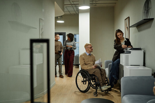 Caucasian young adult man with disability using wheelchair holding documents near printer while woman operating printer and multiethnic colleagues walking in office hallway